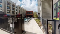 This outdoor pool area is nestled within a contemporary multi-story residential community, offering a resort-inspired retreat surrounded by sleek modern architecture. The pool itself is a standout centerpiece, featuring built-in water fountains that add both visual interest and a soothing ambiance. The surrounding courtyard is thoughtfully designed with a mix of hardscaped pathways and lush green lawn areas, providing a variety of spaces for relaxation and socializing.

Lounge seating and shaded dining areas furnished with tables, chairs, and large umbrellas are positioned throughout the space, offering both sun and shade options for residents. Tall, dark planters filled with vibrant seasonal foliage add striking pops of color and help define the outdoor zones. The area is secured by a gated perimeter with clearly posted safety information, including pool hours and emergency access details.

A brick-accented clubhouse building anchors the far end of the courtyard, featuring floor-to-ceiling glass entry doors that open to interior amenity spaces. The overall design blends industrial and modern aesthetics through the use of mixed materials including brick, metal fencing, and light-toned concrete pavers. The open-sky setting allows for abundant natural light throughout the day, creating a bright and inviting outdoor environment for residents to enjoy.