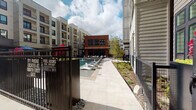 This outdoor pool area is nestled within a contemporary multi-story residential community, offering a resort-inspired retreat surrounded by sleek modern architecture. The pool itself is a standout centerpiece, featuring built-in water fountains that add both visual interest and a soothing ambiance. The surrounding courtyard is thoughtfully designed with a mix of hardscaped pathways and lush green lawn areas, providing a variety of spaces for relaxation and socializing.

Lounge seating and shaded dining areas furnished with tables, chairs, and large umbrellas are positioned throughout the space, offering both sun and shade options for residents. Tall, dark planters filled with vibrant seasonal foliage add striking pops of color and help define the outdoor zones. The area is secured by a gated perimeter with clearly posted safety information, including pool hours and emergency access details.

A brick-accented clubhouse building anchors the far end of the courtyard, featuring floor-to-ceiling glass entry doors that open to interior amenity spaces. The overall design blends industrial and modern aesthetics through the use of mixed materials including brick, metal fencing, and light-toned concrete pavers. The open-sky setting allows for abundant natural light throughout the day, creating a bright and inviting outdoor environment for residents to enjoy.