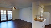 This single-story space presents an open-concept layout that seamlessly integrates the living and dining areas with the adjacent kitchen. Dark hardwood-style flooring runs throughout, offering a warm contrast against the bright white walls and white-painted baseboards. The textured ceiling adds subtle visual depth to the space.

The living area is equipped with a ceiling fan with an integrated light fixture, providing both ventilation and ambient lighting. Natural light filters in through a window with blinds, contributing to the overall brightness of the space. An interior door leads to an additional room, illuminated from within and featuring a bi-fold closet door, suggesting a bedroom or utility space.

The dining area is defined by a pendant light fixture that hangs centrally, creating a natural gathering point. Adjacent to the living and dining areas, the kitchen features white cabinetry, a stone-patterned countertop, and a white range. The kitchen is partially separated by a half-wall partition, maintaining an open feel while providing a subtle visual distinction between the cooking and living spaces.