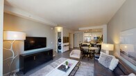 Located at Morgan Parc in Mineola, New York, this single-floor residence offers a thoughtfully designed open-concept layout that seamlessly connects the living, dining, and kitchen areas. The living room is anchored by a large wall-mounted television, a substantial dark media console, and a generously sized sofa, all set against neutral gray walls that create a calm, contemporary atmosphere. A marble-topped coffee table and accent chairs complete the space.

The kitchen is a standout feature, boasting crisp white cabinetry extending to the ceiling, stainless steel appliances including a French-door refrigerator and professional-style range, and a quartz-topped island with an undermount sink illuminated by a sleek pendant light. The island flows naturally into a dedicated dining area.

The bedroom is bathed in natural light from oversized windows and includes a well-appointed closet. Gold-toned sconce lighting adds warmth to the neutral palette. A dedicated study or flex room features a wall-mounted desk, open shelving, and ample space for a multifunctional setup.

The bathroom presents marble-patterned herringbone tile walls, a backlit vanity mirror, a warm wood vanity cabinet, and a soaking tub. An in-unit stacked washer and dryer adds convenience. The overall ambiance is polished, modern, and move-in ready.