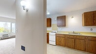 This single-floor unit offers a straightforward, functional layout designed for practical everyday living. The kitchen is equipped with warm oak cabinetry providing ample upper and lower storage, yellow laminate countertops, a white gas range, and a single-basin sink. Light tile flooring runs throughout the kitchen, and wall-mounted lighting fixtures illuminate the space. The kitchen is accessible via a wide doorway, creating an open connection to the rest of the unit.

The living and bedroom areas are finished with neutral-toned wall-to-wall carpeting and cream-painted walls. A baseboard heating system runs along the perimeter, and a wall-mounted HVAC unit is present in the main living area. A large picture window in the living space allows natural light to fill the room and provides an open outdoor view. Additional bedrooms feature windows with blinds and baseboard heaters, maintaining a consistent, understated aesthetic throughout.

The bathroom includes a wood-toned vanity with a white integrated sink, a mirrored medicine cabinet above, and a wall-mounted towel bar. The textured tile flooring adds a functional, easy-to-maintain surface. Overall, the unit presents a clean, utilitarian character with neutral finishes throughout.