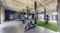 Located at 1693 Broad St in Augusta, Georgia, this expansive single-floor fitness facility offers a generously sized training environment designed with both function and aesthetics in mind. The space showcases an industrial-chic design highlighted by soaring ceilings with exposed ductwork and piping, bold black structural columns, and white painted brick walls that lend a clean, modern character throughout.

The main training floor is divided into distinct functional zones. A central turf area, surfaced with artificial grass, provides dedicated space for functional training and TRX suspension exercises, anchored by a substantial multi-station TRX rig. Surrounding this zone, rubber flooring supports a wide array of strength equipment, including cable machines, selectorized weight stations, free weight benches, and a full dumbbell rack with floor-to-ceiling mirrors reflecting the space.

A cardio section features ellipticals, stationary bikes, treadmills, and a stair climber, positioned near arched double-door entry points and windows that allow natural light to filter in. A glass-panel garage door along one wall adds an airy, open feel and additional natural light. A connecting corridor, accented with decorative wall art and wooden cubbies, provides access between training areas. Linear LED lighting throughout ensures consistent, bright illumination across the entire facility.