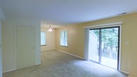 This single-floor residence offers a welcoming and well-lit interior defined by warm, neutral-toned walls and soft carpeted flooring throughout. The main living area is generously proportioned, creating a comfortable and airy atmosphere that lends itself to a variety of furniture arrangements. A ceiling fan with a built-in light fixture is centrally positioned, providing both ventilation and ambient lighting.

Natural light is a defining feature of this space, with multiple windows along the exterior walls drawing in views of surrounding mature trees and greenery. A full-width sliding glass door opens directly onto an outdoor deck, seamlessly connecting the interior living space to the exterior and offering a pleasant indoor-outdoor flow. Vertical blinds on the sliding door allow for adjustable light and privacy control.

The layout includes access to an additional room or hallway through an interior door, suggesting a functional floor plan with clearly defined living zones. White trim and baseboards provide clean contrast against the warm wall tones, contributing to the overall cohesive aesthetic. The combination of abundant natural light, outdoor access, and a tranquil wooded backdrop makes this a comfortable and inviting single-level living space.