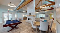 Located in Gulf Shores, Alabama, this single-story Fitness Center and Clubhouse offers a well-appointed community gathering space designed for both relaxation and activity. The centerpiece of the clubhouse is a soaring vaulted great room with exposed natural wood beam trusses, light wood-style flooring, and a dramatic floor-to-ceiling herringbone tile fireplace with a wooden mantel. Circular pendant lights and exposed ductwork add an industrial-coastal aesthetic, while large glass doors invite abundant natural light and views of the surrounding community. The lounge area is furnished with upholstered seating, dining tables, and a billiards table, creating an inviting atmosphere for socializing. A fully equipped community kitchen features a large central island with bar seating, gray cabinetry, stainless steel appliances including a refrigerator and dishwasher, a marble-style tile backsplash, and an overhead range hood. Natural light pours in through oversized windows overlooking the courtyard. The dedicated fitness center is outfitted with rubber flooring and a comprehensive array of cardio and strength equipment, including treadmills, ellipticals, a stationary bike, a rowing machine, and cable machines, supported by mirrored walls and ample ventilation.