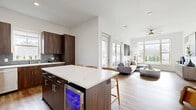 This thoughtfully designed one-bedroom, one-bathroom residence in Olathe, Kansas offers a clean, modern aesthetic across a well-utilized single floor. The entrance hallway with hardwood-style flooring leads seamlessly into the heart of the home. The open-concept kitchen and dining area is a standout feature, showcasing rich dark walnut cabinetry, quartz countertops with a textured tile backsplash, stainless steel appliances, and a spacious center island with bar seating and a built-in beverage cooler — ideal for both cooking and entertaining. The adjoining living room is bright and inviting, filled with natural light from large windows that frame outdoor greenery, and is finished with warm hardwood-style flooring and a ceiling fan. The bedroom offers a comfortable retreat with plush carpeting, a generous window with exterior views, and access to two well-organized closets with built-in shelving. The full bathroom features a wide quartz-topped vanity with dark wood cabinetry, a large mirror, and a glass-enclosed walk-in shower. Additional spaces include a dedicated laundry room and utility room, completing this efficiently designed and stylish residence.