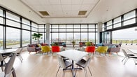 This contemporary amenity floor delivers an exceptional experience through thoughtful design and an abundance of natural light. Anchored by dramatic floor-to-ceiling curtain wall windows that wrap the perimeter, the interior lounge area offers sweeping panoramic views of the surrounding cityscape and a distant body of water. Exposed concrete columns punctuate the open-plan layout, lending an industrial-modern aesthetic that complements the light hardwood flooring throughout.

The lounge zone is furnished with vibrant mid-century modern seating — including bold orange and yellow accent chairs, gray sofas, and colorful area rugs — creating an energetic yet comfortable social environment. A kitchenette and bar area features warm wood cabinetry, subway tile backsplash, built-in flat-screen televisions, and counter-height seating at a sleek black pedestal table, ideal for casual gatherings or events.

Large bi-fold glass doors seamlessly connect the interior to an expansive outdoor terrace. The terrace is finished with large-format pavers and features a pergola-covered seating area, an outdoor fireplace, and multiple dining and lounge groupings — all oriented to maximize the stunning water and skyline views. Planters with seasonal flowers add color to the outdoor space, completing this well-appointed rooftop amenity level.