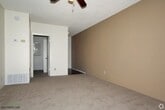 This single-floor residence offers a practical and comfortable layout designed for easy everyday living. The main living area is generously proportioned, featuring soft wall-to-wall carpeting that adds warmth underfoot and a relaxed, welcoming ambiance. A ceiling fan is centrally mounted, providing year-round comfort and air circulation throughout the space. The textured ceiling adds a classic residential character to the room.

Natural light filters in through windows visible toward the back of the space, lending a bright and airy feel to the interior. HVAC vents are thoughtfully positioned throughout, ensuring consistent climate control across the floor plan.

The living area opens seamlessly toward the kitchen, which is partially visible and features dark cabinetry, creating a pleasant contrast against the light-toned walls. A connecting hallway leads further into the home, suggesting additional rooms beyond the main living space.

The overall color palette — soft neutral walls paired with light carpeting — creates a clean, understated aesthetic that is both versatile and easy to personalize. The single-floor layout promotes convenient, step-free accessibility throughout, making this a highly functional and welcoming residential space.