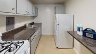 This single-floor unit is defined by its distinctive parquet wood flooring, which extends throughout the living areas and bedrooms, adding warmth and classic character to the space. The flooring features a polished checkerboard-style pattern that reflects natural light effectively.

The living area is generously sized, equipped with a ceiling fan, and anchored by a large sliding glass door that opens to a private balcony. The balcony overlooks lush greenery, providing a pleasant outdoor view and allowing abundant natural light to fill the interior. Structural beams add subtle architectural detail to the ceiling.

Additional rooms, likely used as bedrooms, each feature windows fitted with horizontal blinds, allowing for both natural light and privacy. The neutral white walls throughout provide a clean, versatile backdrop adaptable to various furnishing styles.

The galley-style kitchen is efficiently designed with gray cabinetry, dark granite-style countertops, and light tile flooring. It includes a white refrigerator, a gas range, and a stainless steel sink. Overhead and base cabinet storage is plentiful, maximizing functionality within the compact layout.

Overall, the space maintains a clean, neutral aesthetic with a consistent design language, offering a well-organized and livable environment with practical features throughout.