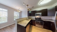 This two-floor residence offers a well-designed layout with a thoughtful mix of modern finishes and functional living spaces. The kitchen serves as a central highlight, showcasing rich dark cabinetry, light granite countertops, and a full suite of stainless steel appliances including a gas range, over-the-range microwave, and refrigerator. A peninsula with an integrated sink and additional counter space adds both utility and an open connection to the adjacent dining and living areas. Track lighting and a pendant chandelier illuminate the space, while wood-look flooring adds warmth throughout the kitchen and main living areas. Natural light filters through slatted blinds on multiple windows, brightening the neutral gray-toned rooms. The carpeted bedroom areas offer comfort and quiet, with one room featuring a dramatic vaulted ceiling that enhances the sense of openness. Crown molding in select rooms adds a refined architectural touch. The bathroom is outfitted with a full soaking tub and shower combination, accented by classic white tile with a decorative tan tile border. A dedicated laundry room with a full-size side-by-side washer and dryer completes the practical amenities, making this a comprehensively equipped multi-level residence.