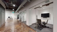 This single-floor office space masterfully combines industrial aesthetics with modern professional design. The layout is anchored by polished concrete floors with rich, variegated tones that run throughout the open corridors and workspaces, creating a striking visual contrast against the clean white walls.

The space features multiple glass-walled conference rooms enclosed by floor-to-ceiling frameless glass partitions and sleek metal hardware, promoting transparency and an open feel while maintaining acoustic separation. Each conference room is equipped with a wall-mounted flat-screen display and a soundbar, complemented by a light wood conference table and ergonomic upholstered chairs.

One conference room includes full-height mirrored wall panels, amplifying the sense of space. A welcoming reception or lounge area features a curved gray sofa, an abstract area rug, a sculptural coffee table, and large-format black-and-white equine artwork, adding a sophisticated artistic touch. Tall tropical plants accent both the lounge and corridor areas.

The open workspace areas showcase exposed ceilings with visible ductwork and industrial pendant lighting, lending an urban loft character. Additional utility rooms and restrooms are accessed via a textured green accent wall corridor. A dedicated workstation area with computers and printing equipment supports day-to-day office operations throughout this thoughtfully designed professional environment.