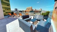 This rooftop terrace is a well-appointed outdoor amenity space designed for relaxation and social gathering, elevated high above a bustling urban skyline. The terrace is finished with large-format porcelain pavers that lend a clean, contemporary aesthetic underfoot, while sleek glass-and-metal railings ensure unobstructed panoramic views of the surrounding cityscape in every direction.

The space is thoughtfully furnished with two distinct seating areas. A sunbathing zone features multiple orange sling-style chaise lounges arranged to maximize sun exposure, while a comfortable lounge area offers modular upholstered seating in cool blue-grey tones, arranged around low coffee tables — ideal for conversation or casual gatherings. Potted topiaries provide subtle greenery throughout.

Architecturally, the rooftop is framed by striking exterior walls clad in bold, multicolored panels in shades of orange, yellow, and green, creating a vivid visual backdrop that contrasts dynamically with the open blue sky. Corrugated metal wall sections add industrial texture, and glass-paned double doors provide easy access to the interior below.

The abundant natural light, open-air layout, and sweeping city views collectively create an energetic yet relaxed atmosphere, making this rooftop terrace a standout communal outdoor destination.