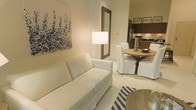 This single-floor space showcases a thoughtfully designed open-concept layout that seamlessly connects the living, dining, and kitchen areas into one cohesive environment. The living area is anchored by a crisp white sofa and complemented by a bold abstract artwork in blue and white tones, adding a touch of artistic character to the neutral walls. A warm-toned floor lamp and a decoratively framed mirror contribute to the inviting ambiance.

The dining area features a rustic round wooden table surrounded by slipcovered white chairs, lending a relaxed yet refined coastal charm. A decorative vase centerpiece adds subtle elegance to the dining setting. Light-toned tile flooring runs throughout, enhancing the sense of openness and reflecting natural light across the space.

The kitchen, visible in the background, is outfitted with stainless steel appliances and warm wood cabinetry, offering a functional yet stylish cooking environment. The overall color palette of whites, beiges, and natural wood tones creates a calm, airy atmosphere throughout. A navy and white striped area rug introduces a nautical accent, tying the coastal aesthetic together cohesively across the entire living space.