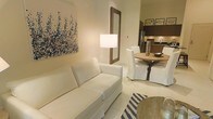 This single-floor space showcases a thoughtfully designed open-concept layout that seamlessly connects the living, dining, and kitchen areas into one cohesive environment. The living area is anchored by a crisp white sofa and complemented by a bold abstract artwork in blue and white tones, adding a touch of artistic character to the neutral walls. A warm-toned floor lamp and a decoratively framed mirror contribute to the inviting ambiance.

The dining area features a rustic round wooden table surrounded by slipcovered white chairs, lending a relaxed yet refined coastal charm. A decorative vase centerpiece adds subtle elegance to the dining setting. Light-toned tile flooring runs throughout, enhancing the sense of openness and reflecting natural light across the space.

The kitchen, visible in the background, is outfitted with stainless steel appliances and warm wood cabinetry, offering a functional yet stylish cooking environment. The overall color palette of whites, beiges, and natural wood tones creates a calm, airy atmosphere throughout. A navy and white striped area rug introduces a nautical accent, tying the coastal aesthetic together cohesively across the entire living space.