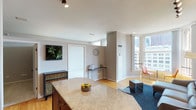 This single-floor urban residence offers a well-designed open-concept layout that seamlessly connects the kitchen, dining, and living areas. The kitchen is outfitted with rich cherry wood cabinetry, granite countertops, a full suite of black appliances including a refrigerator, electric range, built-in microwave, and dishwasher, along with a central island that provides additional prep and gathering space. Hardwood flooring flows throughout the main living areas, complementing the neutral gray walls and clean architectural lines.

The living area benefits from generous natural light streaming through large windows that frame urban cityscape views, creating a bright and inviting atmosphere. Track lighting overhead provides adjustable illumination throughout. A dedicated dining nook positioned near the windows offers an ideal spot for casual meals with a city backdrop.

The bathroom is finished in a crisp, neutral palette featuring tile flooring, a full-size tub and shower combination with safety grab bars, a white vanity with ample cabinet storage, and a mirror. A built-in linen closet adjacent to the bathroom provides practical storage. The overall ambiance of the space is modern, clean, and urban.