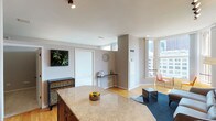 This single-floor urban residence offers a well-designed open-concept layout that seamlessly connects the kitchen, dining, and living areas. The kitchen is outfitted with rich cherry wood cabinetry, granite countertops, a full suite of black appliances including a refrigerator, electric range, built-in microwave, and dishwasher, along with a central island that provides additional prep and gathering space. Hardwood flooring flows throughout the main living areas, complementing the neutral gray walls and clean architectural lines.

The living area benefits from generous natural light streaming through large windows that frame urban cityscape views, creating a bright and inviting atmosphere. Track lighting overhead provides adjustable illumination throughout. A dedicated dining nook positioned near the windows offers an ideal spot for casual meals with a city backdrop.

The bathroom is finished in a crisp, neutral palette featuring tile flooring, a full-size tub and shower combination with safety grab bars, a white vanity with ample cabinet storage, and a mirror. A built-in linen closet adjacent to the bathroom provides practical storage. The overall ambiance of the space is modern, clean, and urban.