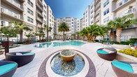 Located at 1305 Morgan Stanley Ave in Winter Park, Florida, this beautifully designed outdoor amenity courtyard delivers a resort-like atmosphere at the heart of a modern multi-story residential community. The centerpiece of the space is a generously sized, curved swimming pool with crystal-clear turquoise water, flanked by sun loungers and surrounded by lush tropical landscaping. Towering palms and clusters of tropical trees are thoughtfully placed throughout, creating natural shade and a vibrant, verdant ambiance beneath the Florida sun. A decorative stone water fountain serves as a striking focal point near the pool's edge, adding an elegant touch to the hardscaped surroundings. A wooden pergola structure anchors one end of the courtyard, outfitted with comfortable wicker seating upholstered in teal and featuring overhead string lighting for evening gatherings. Tucked along garden pathways, canopied daybed swings offer intimate relaxation spots framed by manicured hedges and colorful tropical plantings. Large ornamental planters, warm-toned mulched garden beds, and patterned paver walkways unify the design throughout. The multi-story building facades enclosing the courtyard on all sides feature balconies that overlook this vibrant outdoor retreat, creating a cohesive and inviting community environment.