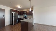 Located at 1386 E 105th St in Cleveland, Ohio, this single-floor residence offers a thoughtfully designed open-concept layout that seamlessly connects the kitchen and living areas. The kitchen is a standout feature, showcasing rich dark espresso cabinetry, granite countertops, and a full suite of stainless steel appliances including a refrigerator, electric range, and over-the-range microwave. A central island with an integrated sink and matching granite surface provides additional prep space and serves as a natural gathering point. Pendant lighting suspended above the island adds a polished, modern touch, while a classic subway tile backsplash lends subtle texture to the cooking area. Wood-style plank flooring extends throughout the main living space, contributing warmth and continuity. The open living area benefits from natural light streaming through a large window, creating a bright and welcoming atmosphere. The bathroom is finished with consistent design elements, featuring a dark wood vanity with a granite countertop, a large mirror, and a combination tub and shower with grab bars. Clean white walls and light flooring keep the bathroom feeling fresh and functional. Overall, the unit presents a cohesive, contemporary aesthetic with quality finishes throughout.