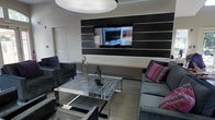 This single-floor space presents a sleek and modern lounge environment designed with a refined contemporary aesthetic. The focal point of the room is a striking horizontal-striped accent wall finished in alternating shades of dark charcoal and metallic grey, which serves as a bold backdrop for a centrally mounted flat-screen television. Subtle backlighting along the base of the accent wall adds a layer of ambient illumination, enhancing the room's polished atmosphere.

The seating arrangement is thoughtfully curated, featuring plush dark grey sofas and armchairs accented with vibrant purple and pink patterned throw pillows, providing a tasteful contrast to the otherwise neutral color palette. A standout marble-topped coffee table with a chrome frame anchors the seating area, adding a touch of elegance and visual weight to the space.

Decorative sculptures displayed on the coffee table contribute artistic character to the room. The flooring is finished in a neutral low-pile carpet that complements the overall grey and charcoal tones throughout. A large, modern circular pendant light overhead provides bright, even illumination, reinforcing the space's clean and contemporary feel. Overall, the lounge exudes a sophisticated, urban ambiance well-suited for social gatherings or relaxed viewing.