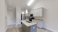 This 1-bedroom, 1-bathroom unit in Autumn Phase II offers a thoughtfully designed single-floor layout with modern finishes throughout. The kitchen serves as a central highlight, featuring a large island with granite countertops and a built-in double sink, making it both a functional workspace and a natural gathering spot. White shaker-style cabinetry lines the walls, providing ample storage, while stainless steel appliances—including a refrigerator, microwave, and range—add a sleek, contemporary touch. Pendant lighting hangs above the island, complementing the flush ceiling fixtures that illuminate the space effectively. The vaulted ceiling in the kitchen area adds an open, airy quality, enhancing the overall sense of spaciousness. Light wood-style flooring runs throughout, contributing to a clean, cohesive aesthetic. The neutral gray and white color palette creates a versatile backdrop that pairs well with a variety of furnishing styles. A door visible near the kitchen suggests convenient access to additional storage or utility space, adding to the unit's practical appeal.