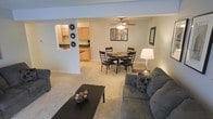 This single-floor residence offers a thoughtfully arranged open-concept layout that seamlessly connects the living, dining, and kitchen areas. The living room is a welcoming space anchored by plush carpeted flooring and furnished with comfortable seating arranged around a dark coffee table, creating an inviting atmosphere for relaxation or entertaining. A ceiling fan overhead provides both practical ventilation and a touch of casual charm. The dining area sits just beyond, accommodating a round four-person dining table beneath a matching ceiling fan with integrated lighting. Decorative wall art adds a warm, personalized touch to the neutral-toned walls throughout. The kitchen is accessible via a pass-through window that maintains visual openness between spaces, featuring light wood cabinetry, dark countertops, and efficient use of the available space. The consistent carpeted flooring throughout the main living areas ties the open floor plan together cohesively. Neutral wall tones contribute to a bright, airy ambiance, while tasteful decorative accents — including wall-mounted sunburst pieces and framed artwork — add character and warmth to the overall environment.