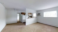 This single-floor residence offers a practical and functional layout with an open feel throughout its main living areas. The living space is finished with wall-to-wall neutral carpet, providing a soft, comfortable underfoot experience. A ceiling fan is centrally mounted overhead, complementing climate control provided by a wall-mounted air conditioning unit and baseboard heating along the exterior wall. Natural light filters in through multiple windows, keeping the space bright and airy.

The kitchen is defined by a half-wall with a breakfast bar-style pass-through counter, creating a visual and functional connection to the living area without fully closing off the space. The kitchen is equipped with medium-toned wood cabinetry, a white refrigerator, microwave, and a tile backsplash, offering a clean and practical cooking environment.

A hallway visible from the main living area leads toward additional rooms, suggesting a well-organized floor plan. The white walls throughout create a neutral, versatile backdrop. Overall, the space presents a straightforward, functional layout suitable for everyday living, with essential amenities thoughtfully integrated into a compact, single-level design.