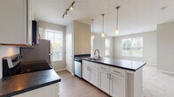 Located at 8352 E Broad St in Blacklick, Ohio, this single-floor residence offers a thoughtfully designed layout with modern finishes throughout. The heart of the home is an open-concept kitchen, which features crisp white cabinetry, dark countertops, a classic subway tile backsplash, and a full suite of stainless steel appliances including a range, microwave, dishwasher, and refrigerator. A large peninsula with an integrated sink and pendant lighting above serves as both a functional workspace and a natural divider between the kitchen and the adjoining living area. Track lighting and multiple windows with white plantation shutters fill the space with ample natural light, creating a bright and welcoming atmosphere. The living area flows seamlessly from the kitchen, offering an open and airy feel with neutral tones and light wood-style flooring. The bedroom is generously proportioned, featuring soft carpeting, a large window overlooking the outdoors, and direct access to a walk-in closet with built-in shelving for ample storage. The bathroom is well-appointed with white cabinetry, a dark countertop vanity, wood-style flooring, and a glass-enclosed walk-in shower equipped with a grab bar, combining practicality with a clean, contemporary aesthetic.