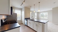 Located at 8352 E Broad St in Blacklick, Ohio, this single-floor residence offers a thoughtfully designed layout with modern finishes throughout. The heart of the home is an open-concept kitchen, which features crisp white cabinetry, dark countertops, a classic subway tile backsplash, and a full suite of stainless steel appliances including a range, microwave, dishwasher, and refrigerator. A large peninsula with an integrated sink and pendant lighting above serves as both a functional workspace and a natural divider between the kitchen and the adjoining living area. Track lighting and multiple windows with white plantation shutters fill the space with ample natural light, creating a bright and welcoming atmosphere. The living area flows seamlessly from the kitchen, offering an open and airy feel with neutral tones and light wood-style flooring. The bedroom is generously proportioned, featuring soft carpeting, a large window overlooking the outdoors, and direct access to a walk-in closet with built-in shelving for ample storage. The bathroom is well-appointed with white cabinetry, a dark countertop vanity, wood-style flooring, and a glass-enclosed walk-in shower equipped with a grab bar, combining practicality with a clean, contemporary aesthetic.