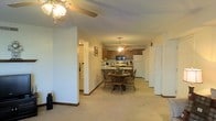 This single-floor residence is designed with an open-concept layout that seamlessly connects the living, dining, and kitchen areas into one cohesive space. The living area is finished with neutral beige carpeting and light-colored walls, creating a warm and understated ambiance. A ceiling fan with a light fixture anchors the foreground of the space, providing both functionality and subtle style.

Moving deeper into the layout, a round wooden dining table with matching chairs sits centrally, illuminated by a pendant ceiling fan above, reinforcing a cozy and practical dining setting. The kitchen, positioned at the rear, features warm medium-toned wood cabinetry and includes standard appliances such as a white refrigerator, microwave, and range. A peninsula counter extends from the kitchen, offering an informal eat-in option that bridges the kitchen and dining areas.

Structural columns punctuate the open floor plan, adding architectural definition without fully separating the zones. A hallway to the left of the living area suggests additional rooms beyond the main living space, while a set of bifold doors to the right likely conceals a storage closet or utility area. The textured ceiling and consistent carpeting throughout contribute to a uniform, comfortable atmosphere.