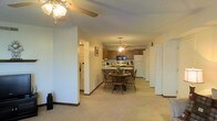 This single-floor residence is designed with an open-concept layout that seamlessly connects the living, dining, and kitchen areas into one cohesive space. The living area is finished with neutral beige carpeting and light-colored walls, creating a warm and understated ambiance. A ceiling fan with a light fixture anchors the foreground of the space, providing both functionality and subtle style.

Moving deeper into the layout, a round wooden dining table with matching chairs sits centrally, illuminated by a pendant ceiling fan above, reinforcing a cozy and practical dining setting. The kitchen, positioned at the rear, features warm medium-toned wood cabinetry and includes standard appliances such as a white refrigerator, microwave, and range. A peninsula counter extends from the kitchen, offering an informal eat-in option that bridges the kitchen and dining areas.

Structural columns punctuate the open floor plan, adding architectural definition without fully separating the zones. A hallway to the left of the living area suggests additional rooms beyond the main living space, while a set of bifold doors to the right likely conceals a storage closet or utility area. The textured ceiling and consistent carpeting throughout contribute to a uniform, comfortable atmosphere.