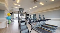 This single-floor fitness center is a comprehensively outfitted facility designed to support a wide range of workout routines. The space is bright and open, featuring light wood-style flooring and neutral tones accented by soft gray and tan wall banding that create a clean, modern aesthetic. Ceiling fans and flush-mounted linear lighting ensure the environment remains comfortable and well-lit throughout.

The cardio zone is anchored by a row of Matrix-brand treadmills, elliptical trainers, stair climbers, and upright cycling bikes, each equipped with individual display screens for entertainment and performance tracking. A dedicated free-weight area houses a full rack of dumbbells alongside a weight bench and mirrors that amplify the sense of space.

A secondary zone offers additional ellipticals, stationary bikes, and a collection of stability and exercise balls mounted on a vertical rack, alongside a standing desk area ideal for fitness consultations or programming. A wall-mounted flat-screen television provides additional entertainment.

One of the most striking architectural features is the full-height glass garage door that opens the space to outdoor views of what appears to be a resort-style pool and landscaped surroundings, flooding the interior with natural light. Large windows on opposing walls further enhance the airy, open atmosphere, making this fitness center both functional and inviting.