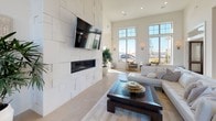 This elegantly designed single-floor communal space blends modern sophistication with warm, inviting details throughout. The grand entryway welcomes guests through double glass doors beneath a statement chandelier, setting the tone for the refined interiors beyond.

The expansive main living area commands attention with soaring double-height ceilings and floor-to-ceiling windows that flood the space with abundant natural light. A striking textured white tile accent wall serves as the room's focal point, housing a sleek linear fireplace and a wall-mounted television. Plush white seating, layered rugs, and curated greenery complete the lounge atmosphere.

Adjacent to the lounge, a well-appointed kitchen features floor-to-ceiling white shaker cabinetry, a herringbone tile backsplash, stainless steel appliances, and a generously sized island topped with veined white marble countertops. Gold-toned geometric pendant lights add a luxurious finishing touch, while woven rattan barstools provide casual seating.

The dining area is bathed in natural light from expansive windows that reach toward the ceiling, offering views of an outdoor pool and surrounding landscaping. Elegant wall sconces in brushed gold complement the neutral furnishings, creating a bright and airy atmosphere ideal for gathering. Light-toned large-format tile flooring unifies the entire space with a clean, cohesive aesthetic.