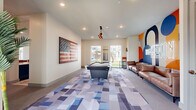 The Gibson is a thoughtfully designed, single-floor amenity space that blends energetic style with functional social areas. The game room serves as a central attraction, featuring a full-size billiards table positioned beneath a striking industrial pendant light cluster. A large, distressed American flag artwork anchors one wall, while the opposite side showcases a bold, vibrant mural bearing "The Gibson" name in vivid orange, navy, and white tones. A leather sofa provides casual seating alongside the mural, complementing the lively atmosphere. Geometric, multi-tone blue and gray carpeting flows throughout, adding texture and visual interest underfoot. Large windows and glass doors fill the space with natural light and offer views of the outdoor courtyard. An adjacent lounge and gathering area provides a more relaxed setting, furnished with blue and tan upholstered armchairs arranged around a round coffee table set with a chess board. A large conference-style table with leather chairs accommodates group gatherings, while circular halo pendant lights overhead lend a modern, polished ambiance. A wall-mounted display screen and additional seating areas make this zone equally suited for both social and productive use. Throughout, the clean white walls and open layout create a bright, welcoming environment.