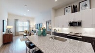 This thoughtfully designed single-floor residence showcases a seamless open-concept layout that connects the kitchen, dining, and living areas into one cohesive, light-filled space. The kitchen is a standout feature, anchored by a generous granite-topped island with an undermount sink, bar seating, and built-in shelving below. Dark cabinetry contrasts elegantly with crisp white upper cabinets, a herringbone tile backsplash, and stainless steel appliances including a French door refrigerator, microwave, and dishwasher. Soaring ceilings throughout lend an airy, expansive feel, while recessed lighting and large windows invite abundant natural light into every corner. The open living area offers generous room for seating and entertaining, flowing effortlessly from the kitchen. The bedroom is a serene retreat, featuring elevated ceilings, wide windows with outdoor views, and pendant lighting. A spacious walk-in closet provides ample storage with dedicated hanging rods and shelving. The full bathroom is well-appointed with a dual-sink granite vanity, dark cabinetry, and a full tub and shower with classic white subway tile surround. Wood-look flooring runs continuously throughout, unifying the home with a warm, contemporary aesthetic.