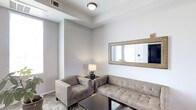 This single-floor space presents a thoughtfully arranged sitting area designed with a clean, contemporary aesthetic. The room is finished in soft, neutral grey tones throughout the walls and ceiling, creating a calm and understated ambiance. A tray ceiling detail adds subtle architectural dimension overhead, complemented by a centrally mounted flush ceiling light that provides steady, even illumination.

The seating arrangement features a tufted sofa and a matching armchair upholstered in taupe-grey fabric, positioned around a dark wood coffee table to create an intimate conversational grouping. A small side table with a warm-toned table lamp sits between the two seating pieces, adding a layer of soft, ambient light to the space.

A large rectangular mirror with a woven gold-toned frame is mounted prominently on the main wall, visually expanding the room and reflecting light to enhance the sense of space. The flooring is finished in a light tile, contributing to the bright, clean feel of the interior. A wall-mounted HVAC unit is visible near the ceiling, ensuring climate control. Overall, the space conveys a polished, functional character suited for comfortable, everyday use.