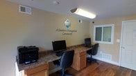 This single-story business center, operated by A Southern Management Community, is a compact and functional workspace designed to serve residents with essential computing needs. The room features warm hardwood flooring that contrasts neatly with the light beige walls, creating an approachable and professional atmosphere. A built-in granite-topped workstation runs along two walls, providing ample desk space and housing multiple computer monitors, a printer, and ergonomic office chairs. Cabinetry beneath the countertop offers additional storage, keeping the space organized and tidy. Overhead fluorescent lighting ensures the room is well-lit, while a window along one wall allows for natural light to supplement the interior fixtures. The branded Southern Management Community logo is prominently displayed on the main wall, reinforcing the professional identity of the space. Ventilation is provided through a ceiling vent, and the overall layout is straightforward and efficient, making it easy for users to navigate. This business center offers a convenient and well-equipped environment for everyday computing tasks.