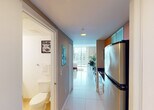 Located at 1750 N Bayshore Dr in Miami's vibrant Edgewater neighborhood, this single-floor studio offers a well-organized open-concept layout flooded with natural light. The main living area is anchored by large floor-to-ceiling windows that frame city views, allowing abundant daylight to reflect off the polished white tile flooring throughout. The sleeping and living zones are thoughtfully combined, featuring a full-sized bed, a dark wood entertainment center with a flat-screen TV, a compact dining table with modern chairs, and lush indoor plants that add a tropical accent. A bold piece of wall art further enhances the contemporary aesthetic. The galley-style kitchen is efficiently designed with warm wood cabinetry featuring frosted glass upper panels, dark granite countertops, a mosaic tile backsplash, stainless steel appliances including a full-sized refrigerator, and a built-in microwave. A narrow hallway connects the kitchen to the main entry and provides access to the bathroom. The bathroom includes a wide vanity with a stone-toned countertop, an undermount sink, a large illuminated mirror with Hollywood-style lighting, and a shower-tub combination. This compact yet complete Miami studio is ideal for those seeking urban convenience in a bright, modern setting.