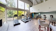 Located in Vestavia Hills, Alabama, this fitness center offers a comprehensive workout facility across a single floor. The space is defined by its dramatic vaulted ceilings with exposed wooden beams, which create an open and airy atmosphere throughout the main exercise area. Expansive floor-to-ceiling windows flood the cardio room with abundant natural light and provide lush, tree-lined outdoor views, enhancing the workout experience.

The main exercise room features light hardwood-style flooring and houses a selection of treadmills, elliptical machines, and stationary bikes, along with a dedicated free weight station equipped with a full rack of dumbbells. A striking spiral staircase anchors one corner of the room, adding an architectural focal point, while a water station is conveniently positioned nearby.

A separate cycling studio is carpeted and lined with spin bikes, wall mirrors, and built-in lockers, offering a dedicated space for group or individual cycling workouts. The lounge area provides a welcoming retreat with a full-height painted brick fireplace, a wall-mounted television, high-top seating, and direct access to an outdoor deck. A hallway connects the various zones of the facility, ensuring smooth flow throughout the space.