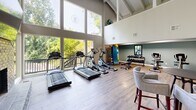 Located in Vestavia Hills, Alabama, this fitness center offers a comprehensive workout facility across a single floor. The space is defined by its dramatic vaulted ceilings with exposed wooden beams, which create an open and airy atmosphere throughout the main exercise area. Expansive floor-to-ceiling windows flood the cardio room with abundant natural light and provide lush, tree-lined outdoor views, enhancing the workout experience.

The main exercise room features light hardwood-style flooring and houses a selection of treadmills, elliptical machines, and stationary bikes, along with a dedicated free weight station equipped with a full rack of dumbbells. A striking spiral staircase anchors one corner of the room, adding an architectural focal point, while a water station is conveniently positioned nearby.

A separate cycling studio is carpeted and lined with spin bikes, wall mirrors, and built-in lockers, offering a dedicated space for group or individual cycling workouts. The lounge area provides a welcoming retreat with a full-height painted brick fireplace, a wall-mounted television, high-top seating, and direct access to an outdoor deck. A hallway connects the various zones of the facility, ensuring smooth flow throughout the space.