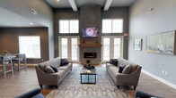 This single-floor space makes a strong architectural impression through its soaring double-height ceilings, accented by exposed white beams that add structural character to the room. The centerpiece of the space is a striking floor-to-ceiling stacked stone fireplace, complete with a warm wood mantel and a wall-mounted television above, creating a commanding focal point that draws the eye upward. Flanking the fireplace are two sets of glass-paned French doors, which allow generous amounts of natural light to flood the interior while providing access to an outdoor area with a wrought-iron railing visible beyond. Additional high clerestory windows further enhance the bright, airy atmosphere. The main seating area is anchored by two substantial gray upholstered sofas arranged symmetrically around a dark-framed coffee table, all resting atop a richly patterned blue area rug. The walls are finished in a sophisticated slate gray tone, complementing the cool-toned décor accented with navy blue throw pillows. The overall ambiance is modern, inviting, and well-appointed, suitable for gathering and relaxation.