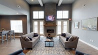 This single-floor space makes a strong architectural impression through its soaring double-height ceilings, accented by exposed white beams that add structural character to the room. The centerpiece of the space is a striking floor-to-ceiling stacked stone fireplace, complete with a warm wood mantel and a wall-mounted television above, creating a commanding focal point that draws the eye upward. Flanking the fireplace are two sets of glass-paned French doors, which allow generous amounts of natural light to flood the interior while providing access to an outdoor area with a wrought-iron railing visible beyond. Additional high clerestory windows further enhance the bright, airy atmosphere. The main seating area is anchored by two substantial gray upholstered sofas arranged symmetrically around a dark-framed coffee table, all resting atop a richly patterned blue area rug. The walls are finished in a sophisticated slate gray tone, complementing the cool-toned décor accented with navy blue throw pillows. The overall ambiance is modern, inviting, and well-appointed, suitable for gathering and relaxation.