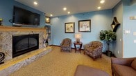 This single-floor community room is thoughtfully designed to accommodate a variety of social activities, offering multiple distinct zones within one expansive, open layout. The walls are painted in a calming steel blue throughout, lending the entire space a cohesive and welcoming ambiance.

The lounge area features comfortable upholstered armchairs in patterned fabric, arranged around side tables and anchored by a stone-surround fireplace with a built-in television and shelving above. Framed botanical artwork and decorative mirrors accent the walls, while recessed lighting and table lamps provide warm illumination.

An adjoining reading nook includes additional seating, a built-in bookshelf stocked with reading materials, and windows overlooking a lush green landscape outside.

The dining area is bathed in natural light from a dramatic bank of tall, grid-paned windows that rise toward a vaulted ceiling, offering views of the surrounding greenery. Round wooden dining tables paired with cushioned chairs comfortably seat multiple guests.

A fully equipped kitchen supports event hosting with wood cabinetry, a full-size refrigerator, electric range, microwave, dishwasher, and a double sink beneath a window with outdoor views. Patterned carpet flows through the main areas, while the kitchen features resilient flooring.