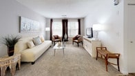 Located at 1312 Town Walk Dr in Hamden, Connecticut, this single-floor residence offers a well-organized layout with a warm, contemporary aesthetic throughout. The living room is a comfortable, carpeted space filled with natural light streaming through large windows dressed with layered blinds and rich drapes, creating an inviting atmosphere for relaxing or entertaining. Adjacent to the living area, a dedicated dining space features chair-rail molding, a pendant light fixture, and carpeted flooring, lending the room a polished, intimate feel. The kitchen is outfitted with white cabinetry, sleek brushed-nickel hardware, light quartz countertops, and stainless steel appliances, with wood-look vinyl plank flooring extending into a convenient laundry area just off the kitchen. The primary bedroom is generously sized with plush carpeting and sliding glass door access to an outdoor area, offering pleasant views of the surrounding greenery. A second bedroom provides flexible use as a guest room or home office, with bright natural light from a large window. The bathroom is clean and modern, featuring a white vanity with ample storage, a well-lit mirror, and wood-look vinyl plank flooring that ties the space together cohesively.