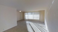 This single-floor space offers a clean, open layout designed for comfortable everyday living. The main living area is a standout feature, characterized by its generous proportions and an abundance of natural light streaming in through a wide bank of windows dressed with vertical blinds. The light filters across the soft, neutral-toned carpeted flooring, creating a warm and inviting atmosphere throughout the day. The walls are finished in a crisp, clean white, lending the space an airy and bright feel that enhances its sense of openness. A flush-mount ceiling light provides additional illumination, complementing the natural daylight. The overall ambiance is calm and understated, with a neutral palette that serves as a versatile backdrop for a variety of interior styles. The layout is straightforward and functional, making efficient use of the available space. The large window wall is a defining architectural element, drawing the eye outward and connecting the interior to the outside environment. This space presents a welcoming and well-lit environment suited for a range of uses.