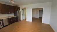 This single-floor residence offers a clean, open layout that connects the kitchen and living areas seamlessly, creating a bright and functional living space. The kitchen is outfitted with dark espresso cabinetry, granite countertops, and stainless steel appliances, including a full-size refrigerator. Track lighting overhead provides focused illumination, complementing the warm hardwood-style flooring that runs throughout the unit. Adjacent to the kitchen, a dedicated laundry nook houses a stacked washer and dryer unit, adding everyday convenience. The open living area flows naturally toward the bedroom, which features a spacious walk-in closet behind a louvered door, offering ample storage. The neutral white walls and clean architectural lines throughout the space contribute to a bright and airy ambiance. The overall design is practical and inviting, making efficient use of the available floor plan while maintaining a comfortable, livable atmosphere.