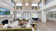 Located at 240 Ivy Meadow Ln in Durham, NC, this single-story community clubhouse offers an expansive and thoughtfully designed gathering space. The centerpiece of the interior is a dramatic living room distinguished by soaring double-height ceilings, an abundance of natural light streaming through oversized clerestory windows, and a stately fireplace framed by built-in cabinetry and display niches. Rich hardwood-style flooring flows throughout, tying the spaces together cohesively. The combined kitchen and living area features sleek countertops, a geometric marble-inspired tile backsplash, a stainless steel range hood, and modern fixtures, seamlessly connecting to a dining area furnished with a round wood table and navy accent chairs. Floor-to-ceiling windows in the dining space offer direct views of the outdoor pool. A dedicated office provides a quiet, functional area, while multiple hallways connect the various zones of the clubhouse with ease. Outdoors, a generously sized resort-style pool is surrounded by lounge chairs, cabana seating, manicured landscaping, and decorative lighting, creating an inviting environment. The grounds are well-maintained with lush greenery, stone walkways, and thoughtfully arranged outdoor seating areas, rounding out this comprehensive community amenity space.