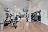 Located at 1400 Chaffee St in Upland, California, this fitness center is a thoughtfully designed exercise facility offering a comprehensive workout environment. The space is defined by soaring vaulted ceilings that create an open, airy atmosphere, complemented by warm hardwood-style flooring throughout.

Natural light streams in through multiple windows positioned along the walls, providing bright and energizing views of the surrounding outdoor area, including a glimpse of a pool. Large floor-to-ceiling mirrors line one full wall, amplifying the sense of space and allowing users to monitor their form during workouts.

The facility is well-stocked with a diverse selection of equipment, including a row of Star Trac treadmills, stationary bikes, a stair climber, and weight benches paired with a full dumbbell rack. Wall-mounted flat-screen televisions are positioned throughout for entertainment during workouts. Ceiling fans ensure comfortable air circulation.

The entrance area features high ceilings, a bulletin board, posted occupancy and facility rules, and a keypad entry system. The clean, white walls and crisp architectural details give the space a modern, polished feel. Direct access to the outdoor pool area is available through a glass-paneled door, making this fitness center a central and convenient amenity.