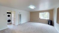 This single-floor space presents a clean, well-maintained interior designed with a neutral and inviting color palette. The main living area is generously proportioned, featuring plush wall-to-wall carpeting in a soft, light gray tone that adds warmth and comfort underfoot. The walls are finished in a combination of crisp white and warm beige, creating a calm and balanced aesthetic throughout.

The ceiling is smooth and white, fitted with a modern flush-mount light fixture that provides even, ambient illumination. A large window along the beige accent wall allows natural light to fill the room while offering views of surrounding greenery, enhancing the sense of openness. White baseboards frame the perimeter of the room, adding a polished, finished appearance.

A tall, vertical wall-mounted heating unit is positioned near the entryway, indicating functional climate control built into the structure. A doorway leading to an adjacent room or hallway is visible, suggesting additional living or utility spaces beyond the main area. Overall, the space conveys a straightforward, functional layout with a light, airy ambiance suited for comfortable everyday living.