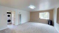 This single-floor space presents a clean, well-maintained interior designed with a neutral and inviting color palette. The main living area is generously proportioned, featuring plush wall-to-wall carpeting in a soft, light gray tone that adds warmth and comfort underfoot. The walls are finished in a combination of crisp white and warm beige, creating a calm and balanced aesthetic throughout.

The ceiling is smooth and white, fitted with a modern flush-mount light fixture that provides even, ambient illumination. A large window along the beige accent wall allows natural light to fill the room while offering views of surrounding greenery, enhancing the sense of openness. White baseboards frame the perimeter of the room, adding a polished, finished appearance.

A tall, vertical wall-mounted heating unit is positioned near the entryway, indicating functional climate control built into the structure. A doorway leading to an adjacent room or hallway is visible, suggesting additional living or utility spaces beyond the main area. Overall, the space conveys a straightforward, functional layout with a light, airy ambiance suited for comfortable everyday living.