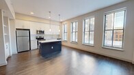 This thoughtfully designed 2-bedroom, 2-bathroom residence features a clean, modern aesthetic carried consistently throughout its single-floor layout. The open-concept living and kitchen area serves as the heart of the home, where rich wood-style flooring, recessed lighting, and a row of tall windows flood the space with natural light and views of the surrounding neighborhood. The kitchen is a standout feature, outfitted with crisp white cabinetry, a white subway tile backsplash, gleaming quartz countertops, and a full suite of stainless steel appliances including a refrigerator, range, microwave, and dishwasher. A navy blue kitchen island with a white countertop provides additional prep space and casual seating, anchored by stylish pendant lighting above. The living area flows seamlessly from the kitchen, creating an inviting and functional shared space. A clean hallway with recessed lighting connects the bedrooms and bathrooms, maintaining a quiet separation from the main living areas. The primary bedroom is a comfortable, well-proportioned retreat filled with natural light from dual windows. The second bedroom features built-in closets with bi-fold doors for practical storage. Both bathrooms are finished with neutral tile, including a glass-enclosed walk-in shower with marble-style tile in one bathroom, paired with modern fixtures throughout.