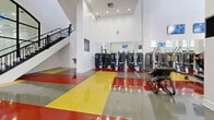 Located at 325 W Adams Blvd in Los Angeles, this two-story fitness facility offers a substantial and thoughtfully designed space dedicated to athletic training and wellness. The larger ground floor serves as the primary exercise area, characterized by its striking polished epoxy flooring in bold red, yellow, and gray zones that define distinct workout areas. Soaring ceilings amplify the sense of openness, while rows of recessed lighting and mounted televisions create a bright, energetic atmosphere throughout. Large arched windows and glass entry doors invite abundant natural light and provide views of the surrounding greenery. A standout rock climbing wall anchors one end of the floor, complemented by extensive strength and cardio equipment, including treadmills, ellipticals, stair climbers, and cable machines. Floor-to-ceiling mirrors line the walls, further enhancing the expansive feel of the space. An ornamental iron staircase connects the ground floor to the upper level, which houses additional cardio equipment along an open-railed mezzanine overlooking the main floor. Supporting spaces include a clean, tiled amenity room accented with iridescent wall finishes and tropical foliage. The facility presents a polished, professional environment suited for high-capacity fitness use.